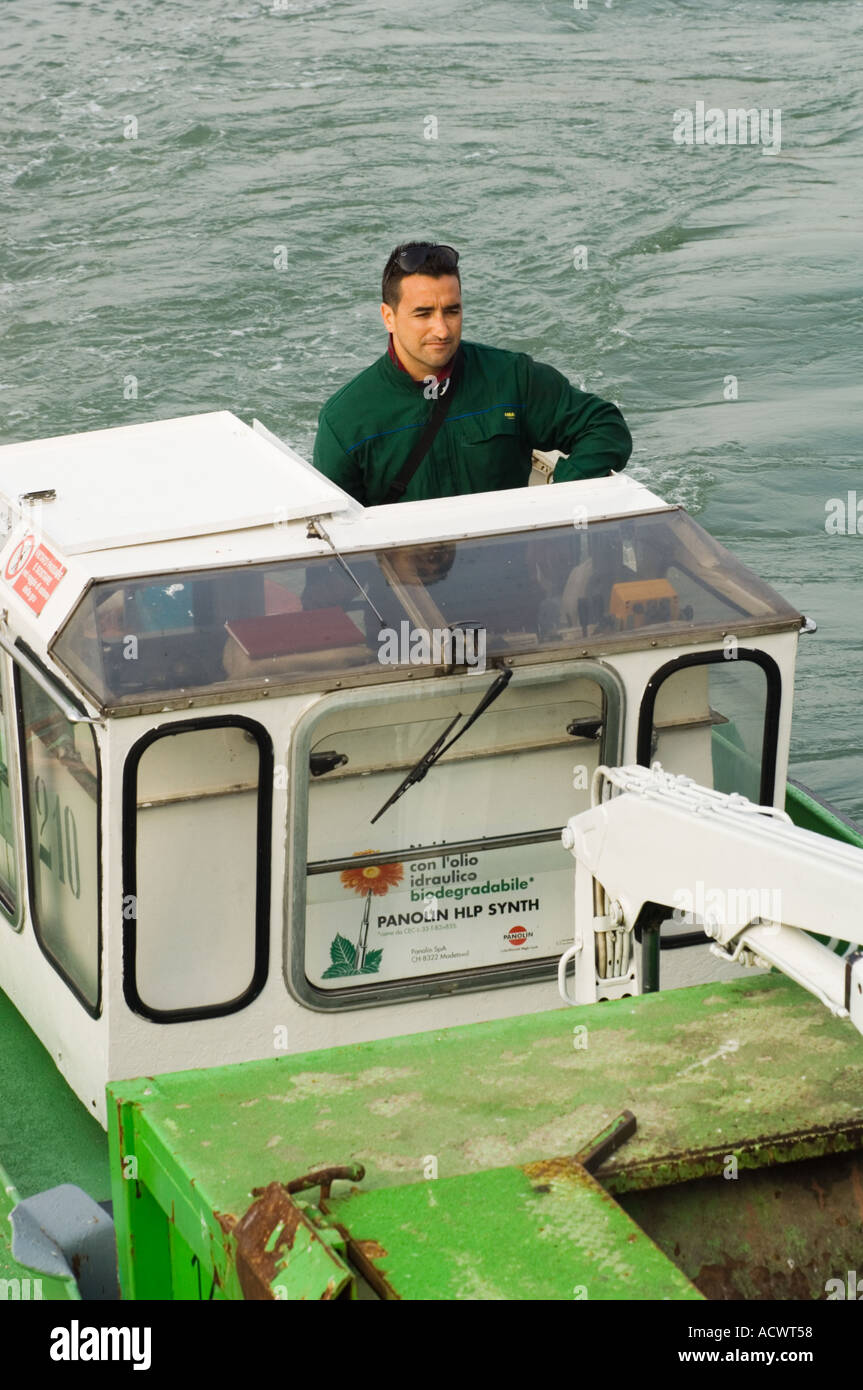 Italian trash man in the pilot house of a green garbage boat in Venice Italy Stock Photo