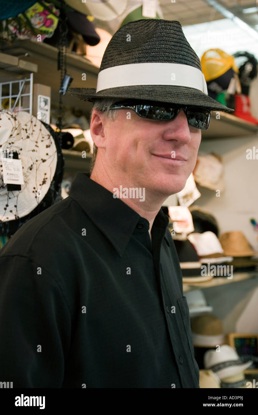 Portrait of a man wearing a black shirt and dark sunglasses trying on a black hat with a white sash in a hat store Stock Photo