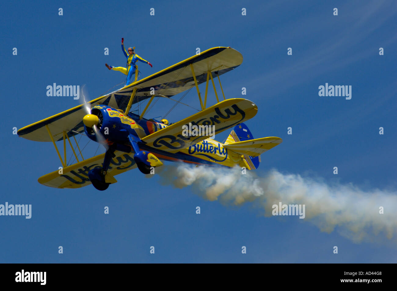 A bi-plane flying in the blue sky with a wing-walker on the top, and a smoke trail trailing behind. Stock Photo