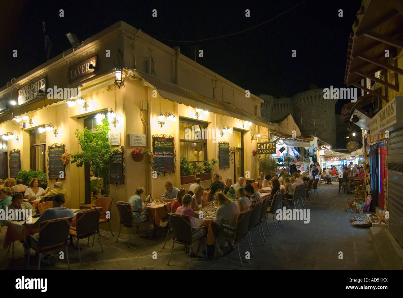 Horizontal wide angle of a traditional Greek taverna with tables and chairs spilling out onto the pavement at night. Stock Photo