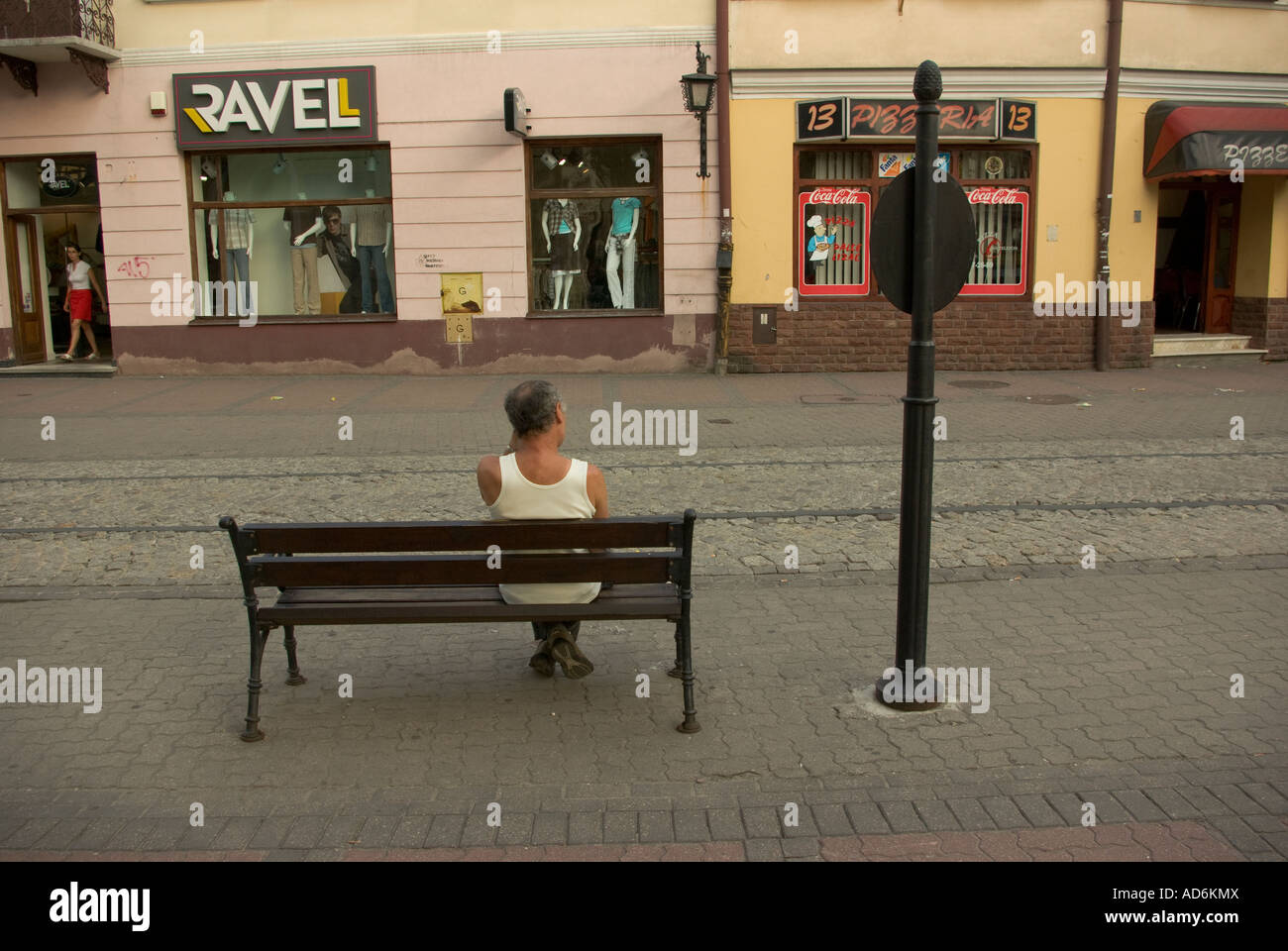 Man in tee-shirt with back to camera sitting on public bench across from stores. Stock Photo