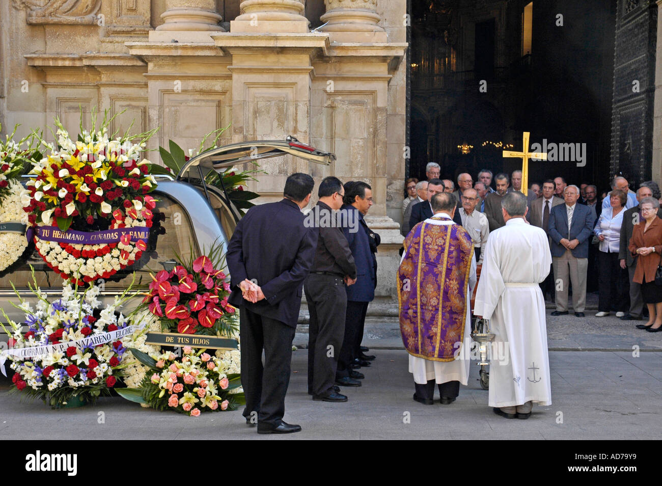 Priest with golden cross and fuenral guests coming out of the church at a funeral, hearse with mourning wreaths Stock Photo