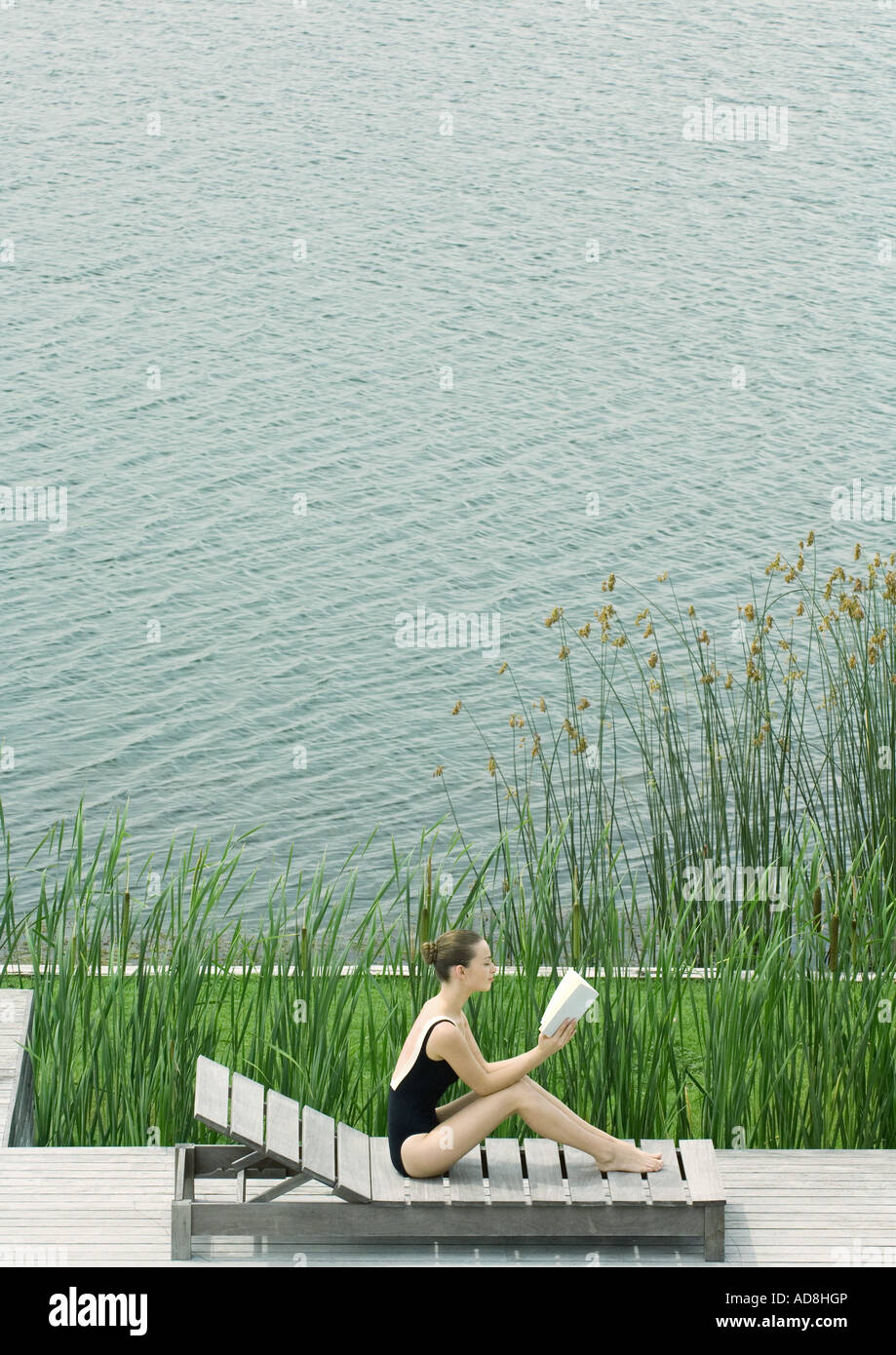 Woman sitting up on lounge chair, reading, next to lake Stock Photo
