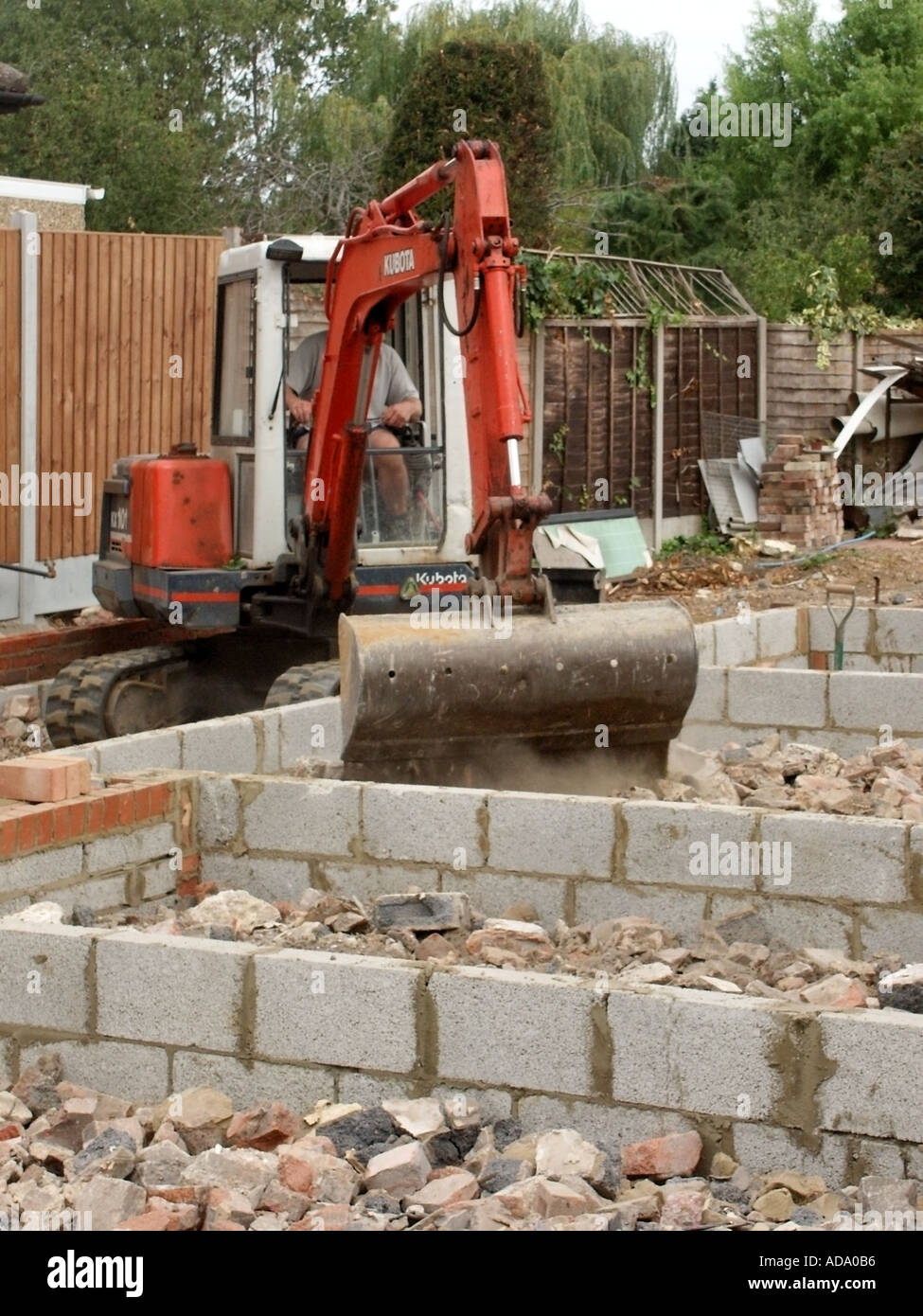 Mini Digger hydraulic excavator & driver placing hardcore rubble between concrete block walls to new house substructure ready for concrete floor UK Stock Photo