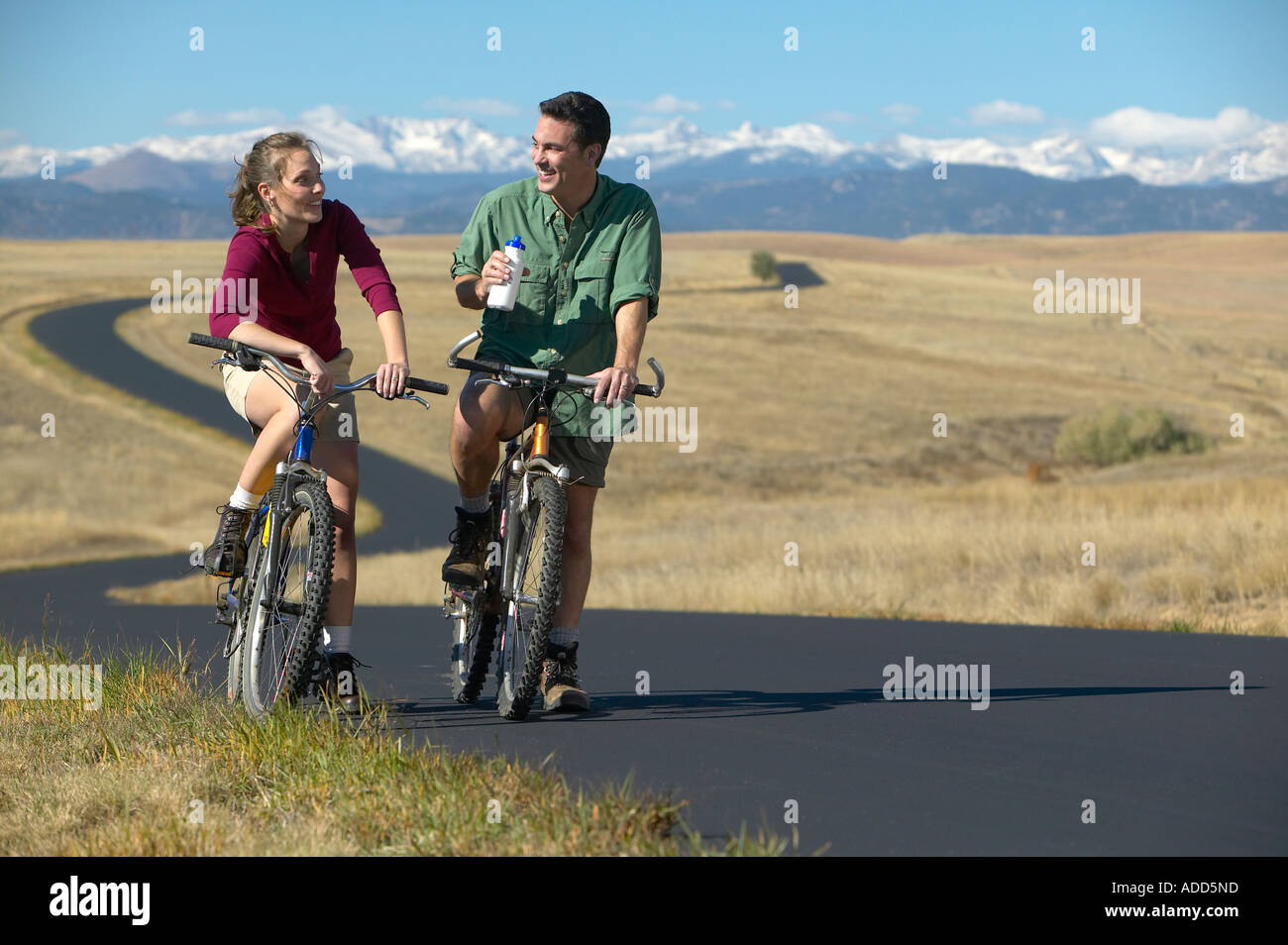 smiling Middle aged couple riding bikes on a trail with mountain range in the background Stock Photo