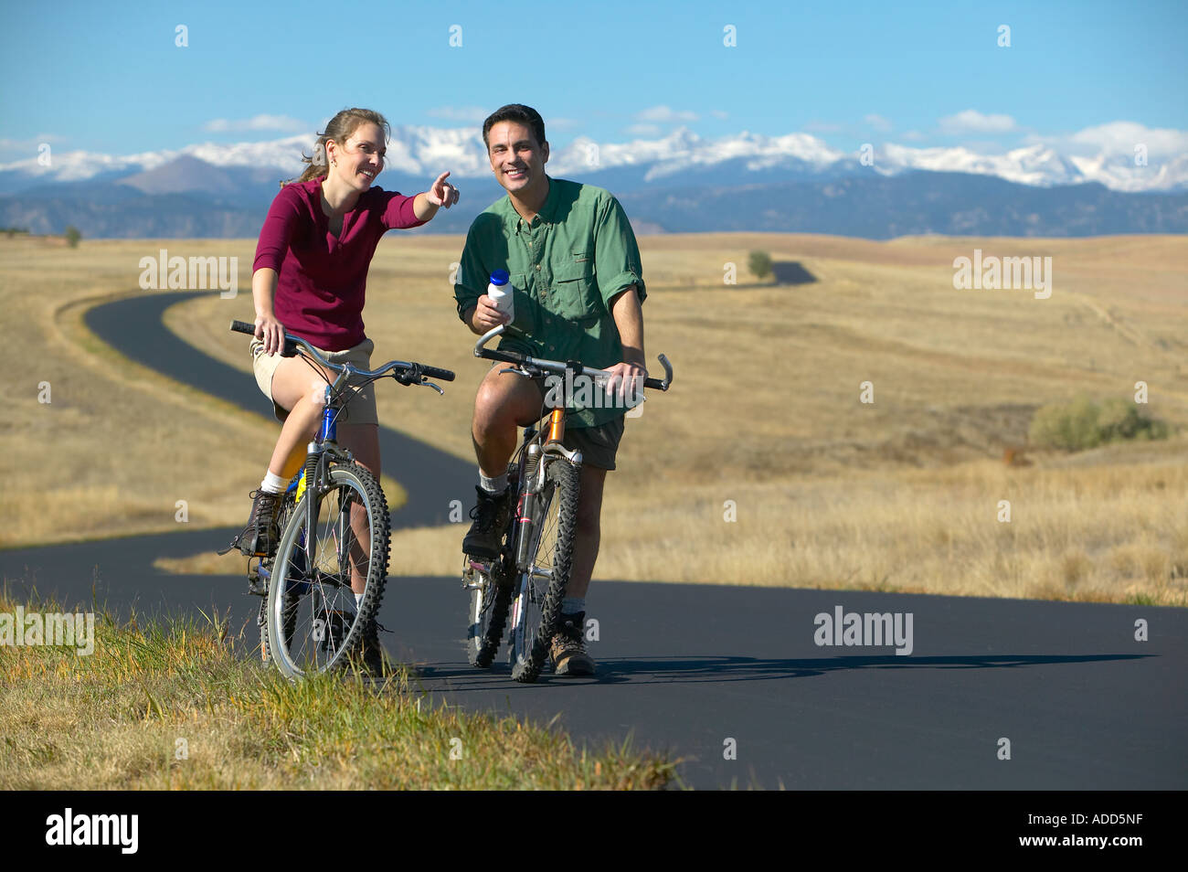 Smiling middle aged couple riding bikes on a trail with mountain range in the background Women points Stock Photo
