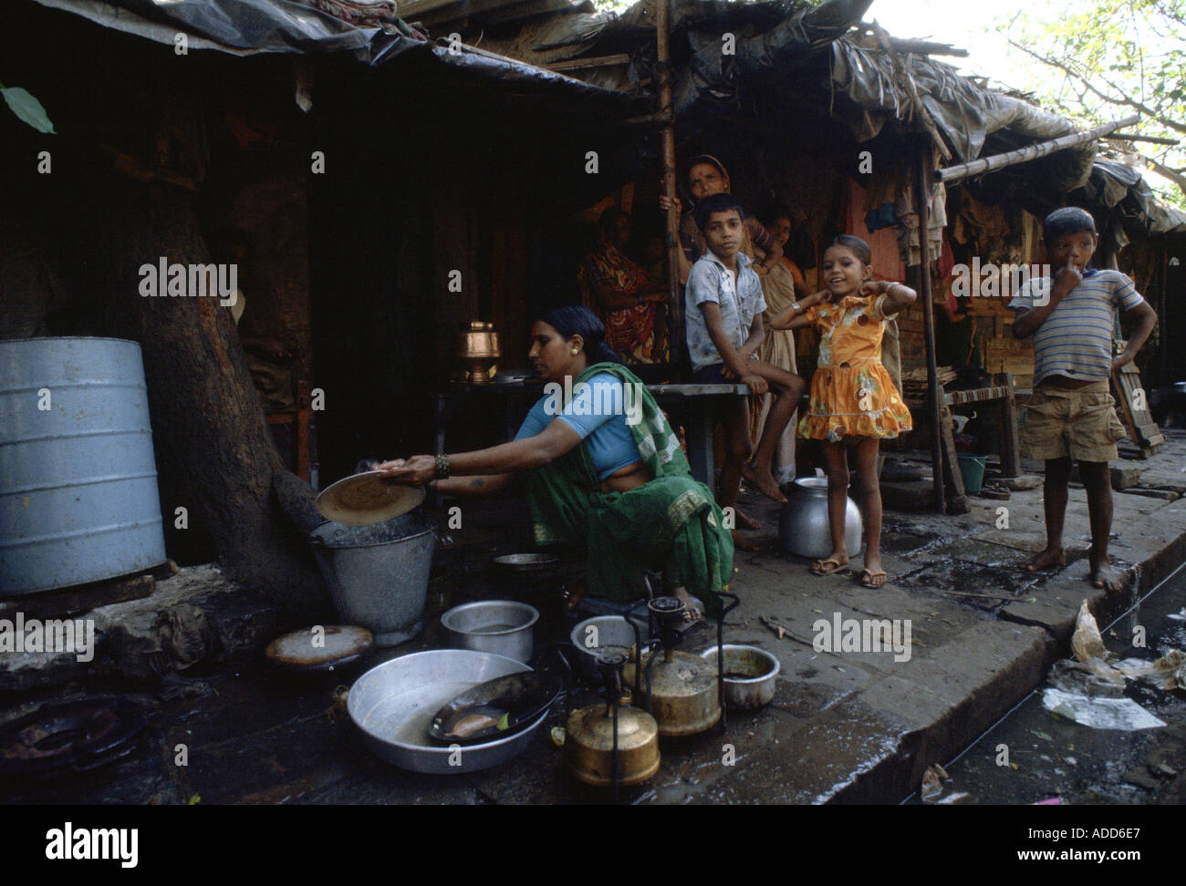 Children watch as woman washes dishes in bucket at shack home on the sidewalk Bombay India Stock Photo