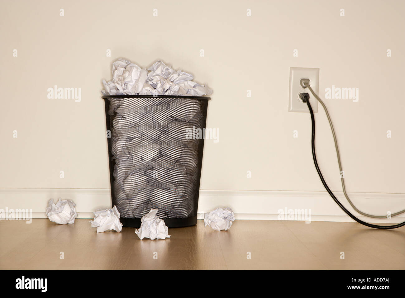 Full wire mesh trash can with crumpled paper next to electrical outlet