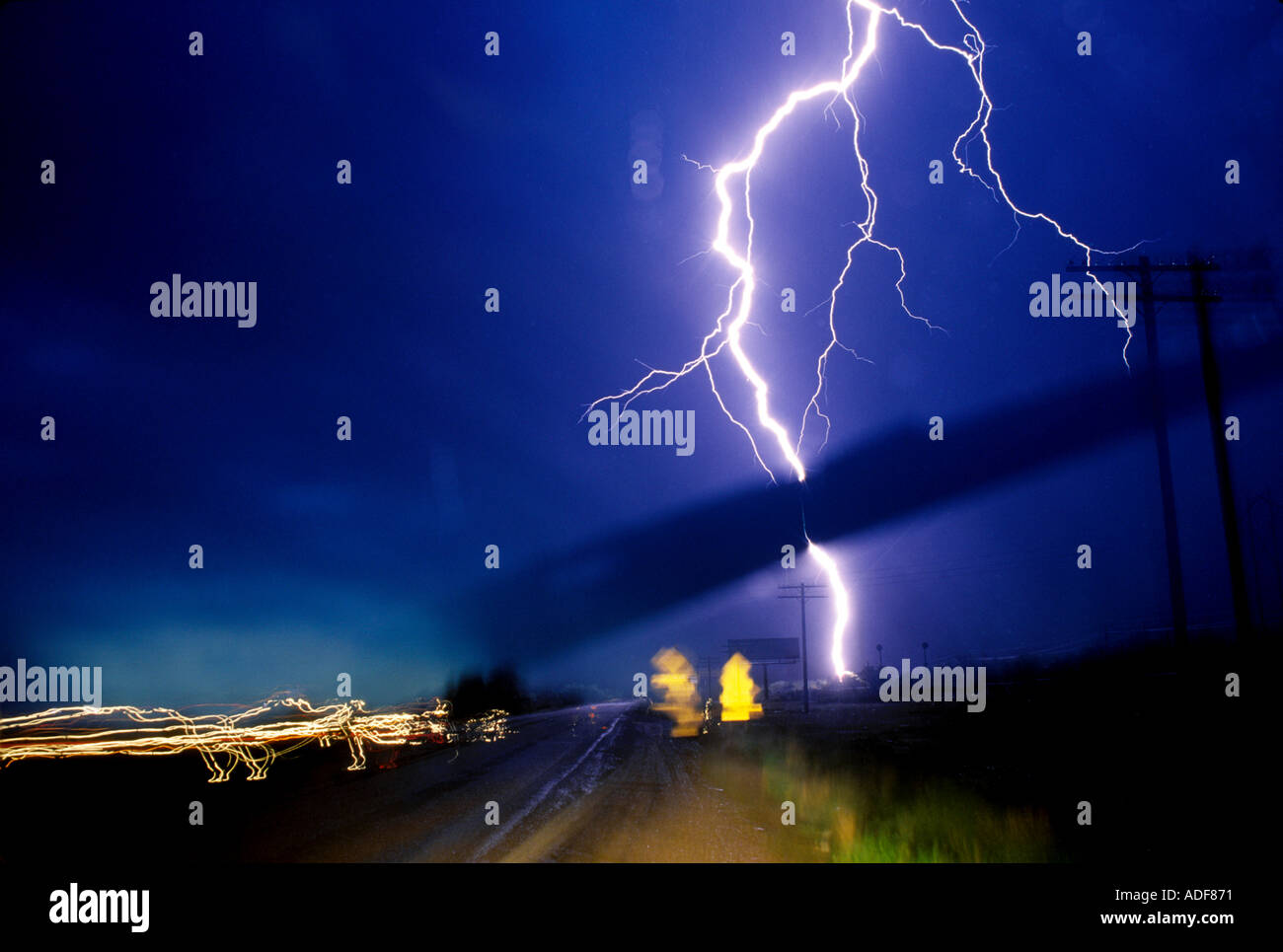 A lightning bolt crashes down next to the highway during a a storm. Lights blur the foreground