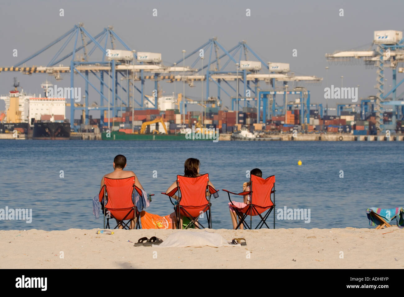 A family on a beach next to a container port at Pretty Bay, Birzebbuga, Malta. Economic development, quality of life, land use and geography. Stock Photo