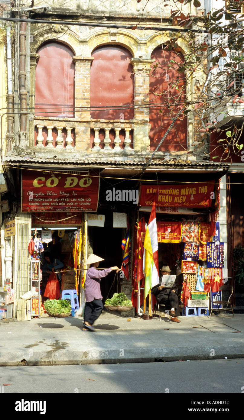 Woman with baskets walking past a shop selling banners and flags on Hang Bong St, Hanoi Old Quarter, Viet Nam Stock Photo