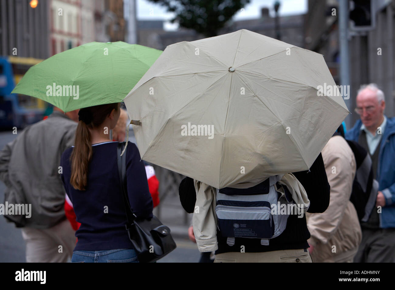 man with backpack and woman with handbag carrying umbrellas waiting to cross the road in dublin Stock Photo