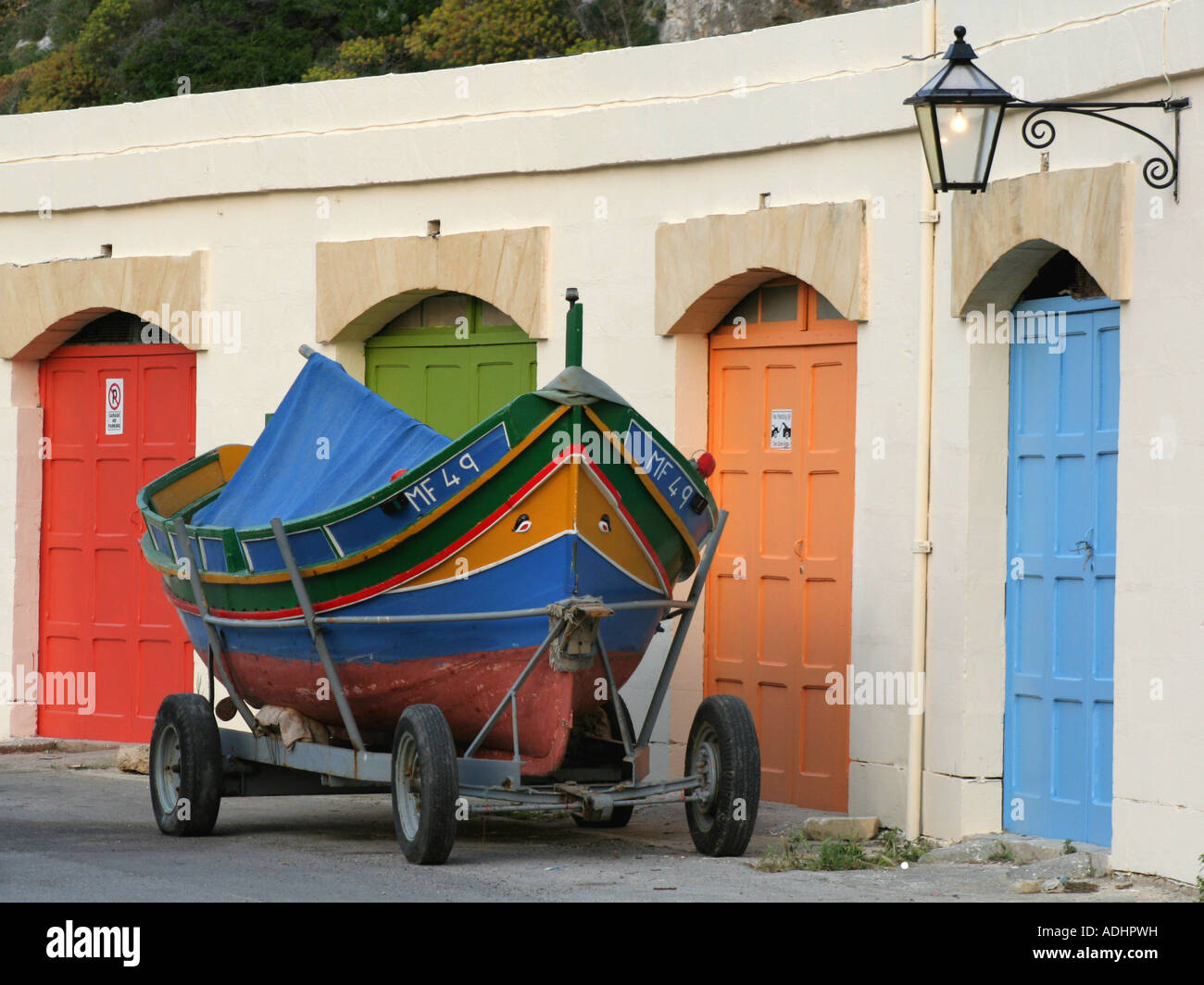 Luzzu (traditional Maltese fishing boat) on a trailer outside a row of garages at Xlendi, Gozo, Malta Stock Photo