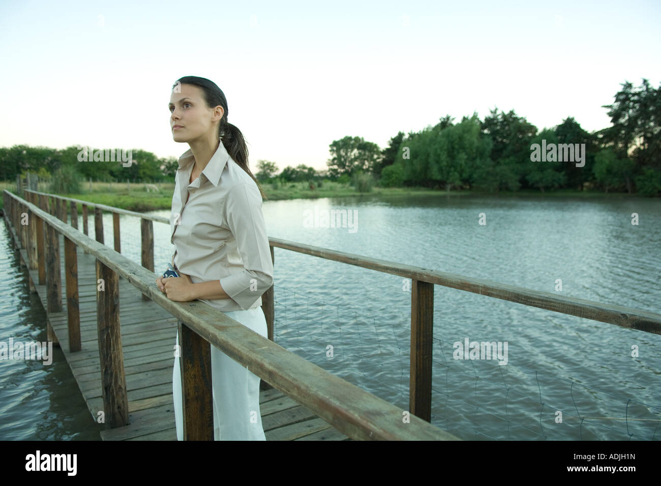 Woman standing on wooden footbridge over lake, looking into distance Stock Photo