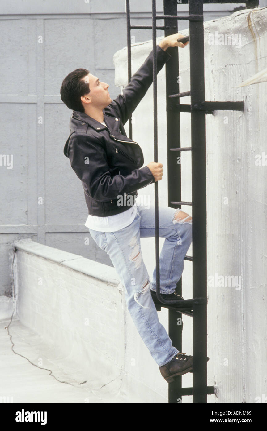 Young Caucasian Man Wearing Leather Motorcycle Jacket Climbing Ladder To Building Roof Stock Photo