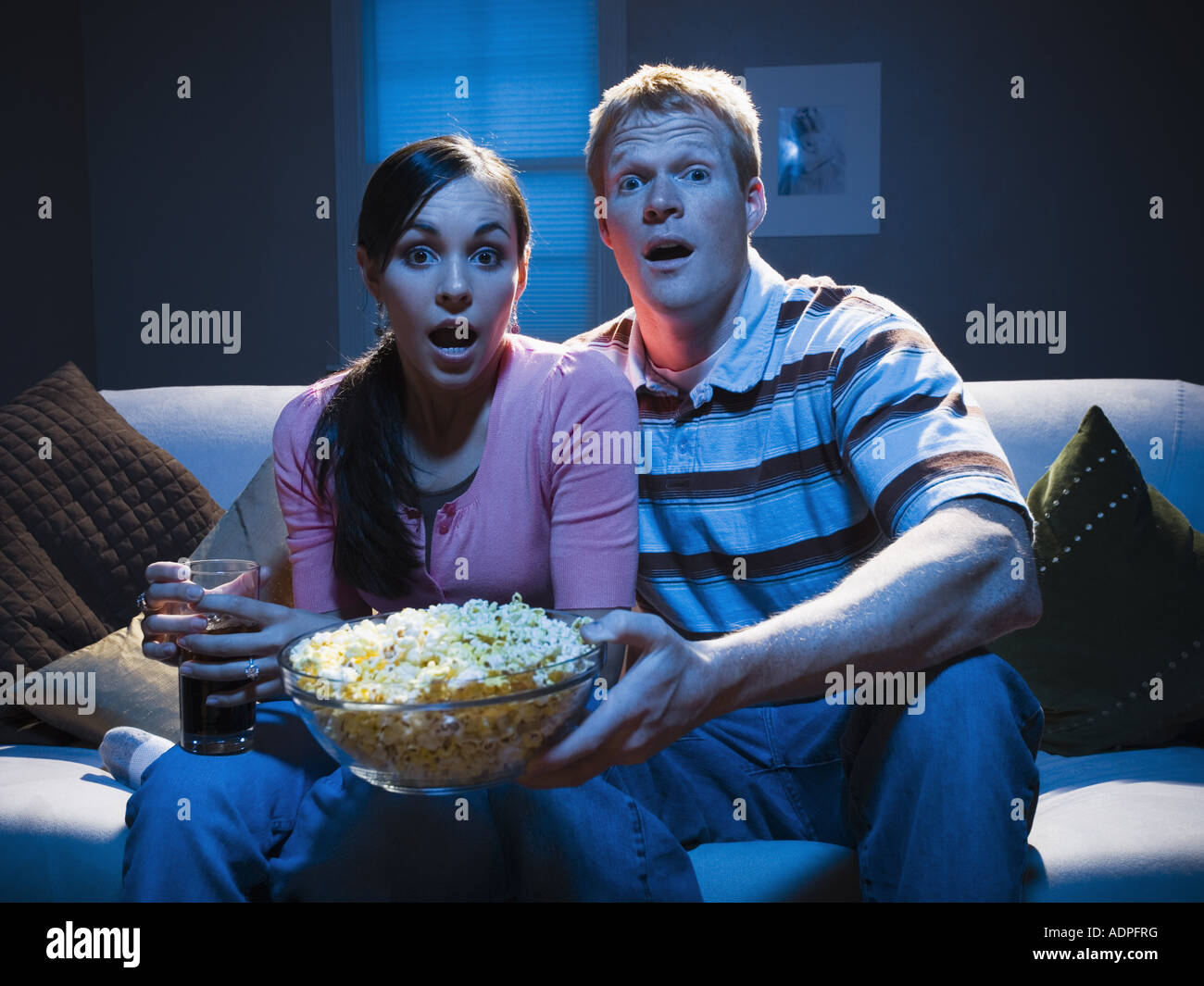 Couple on sofa with bowl of popcorn looking shocked Stock Photo