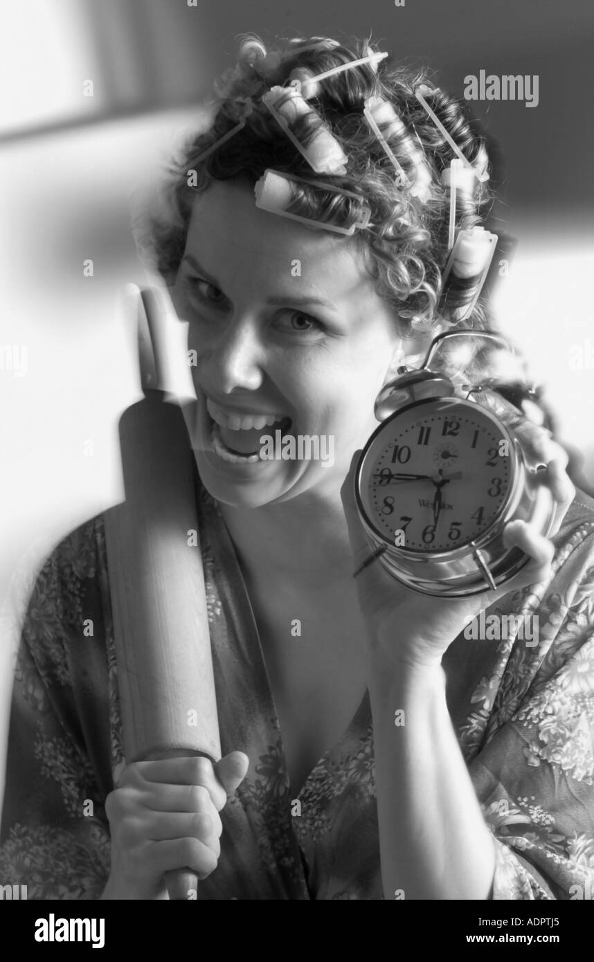 Woman in hair curlers with rolling pin and alarm clock greeting hubby at the door Stock Photo