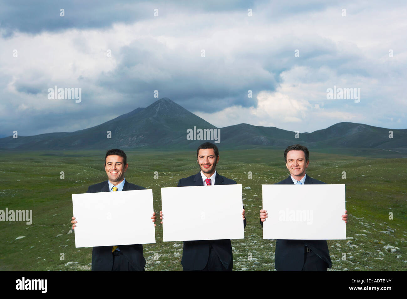 Three smiling businessmen standing in mountain field holding blank ...