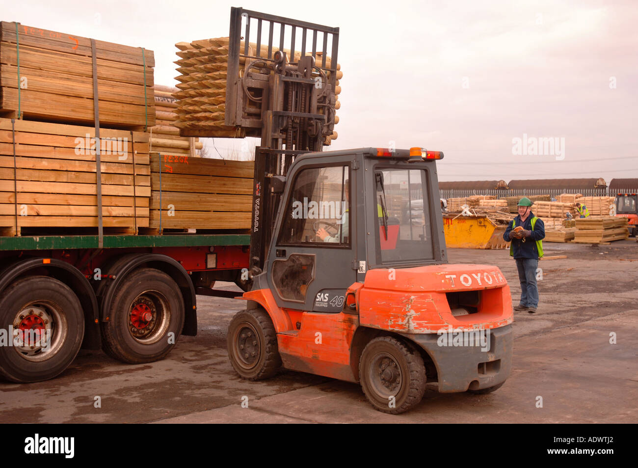 A FORK LIFT TRUCK LIFTS TIMBER FENCING POSTS ONTO AN ARTICULATED LORRY ...