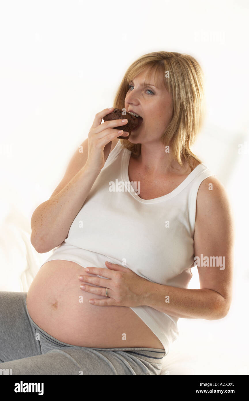 ATTRACTIVE HEALTHY YOUNG PREGNANT WOMAN SITTING ON BED EATING CHOCOLATE MUFFIN Stock Photo