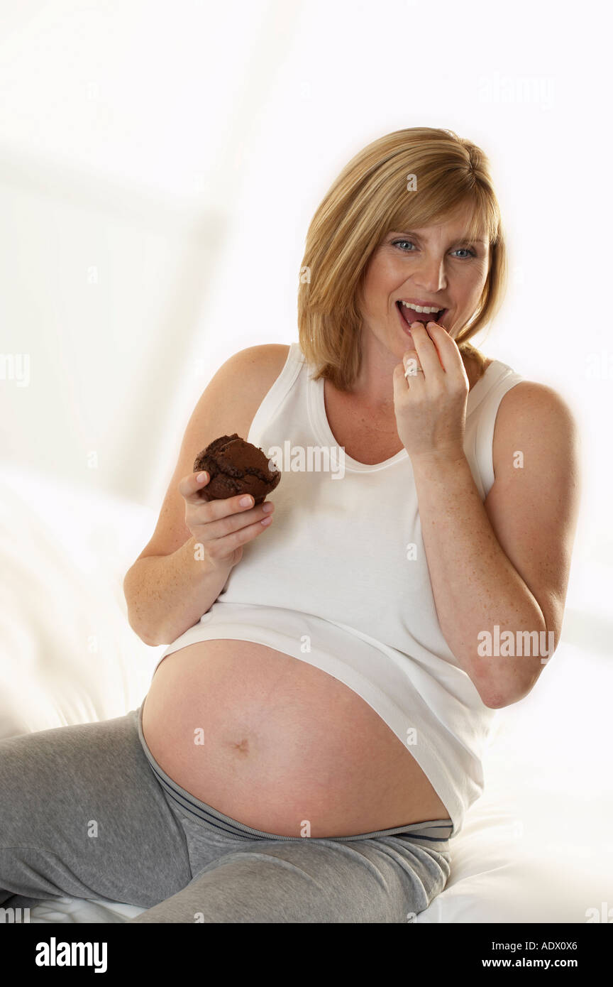 ATTRACTIVE HEALTHY YOUNG PREGNANT WOMAN SITTING ON BED EATING CHOCOLATE MUFFIN Stock Photo