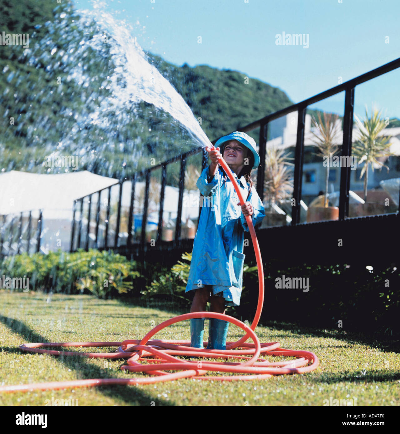 Children young girl 5 10 blue raincoat hose hoses raincoats watering stream jet field sunny childhood holding looking away fence Stock Photo