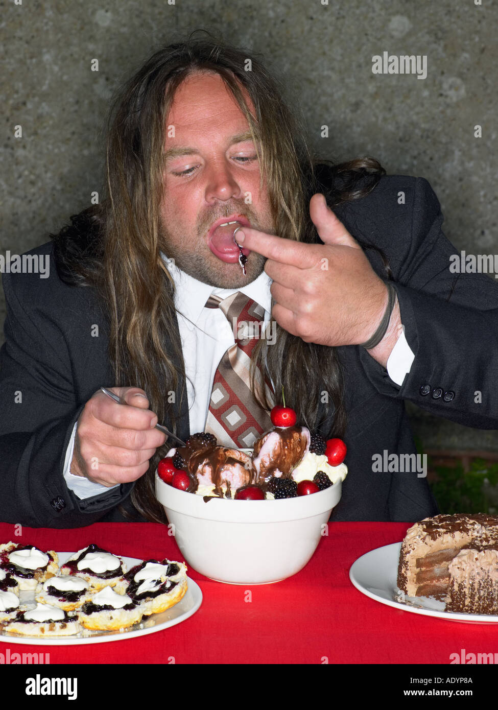 Long haired fat man eating stuffing himself with cake and ice cream Stock Photo