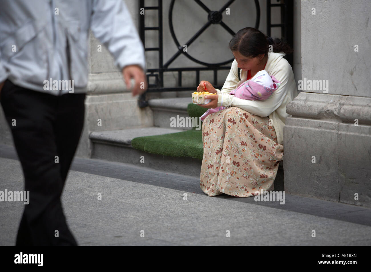 young gypsy beggar woman holding what looks like a baby wrapped in blanket eating chips in doorway as man walks past oconnell street dublin Stock Photo