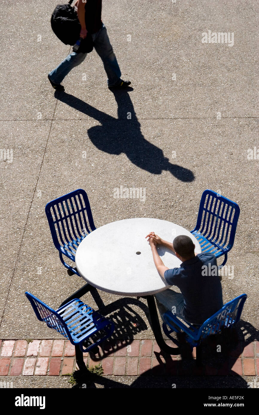 Man sitting waiting at round table in outdoor eating area with shadow of man walking by Stock Photo