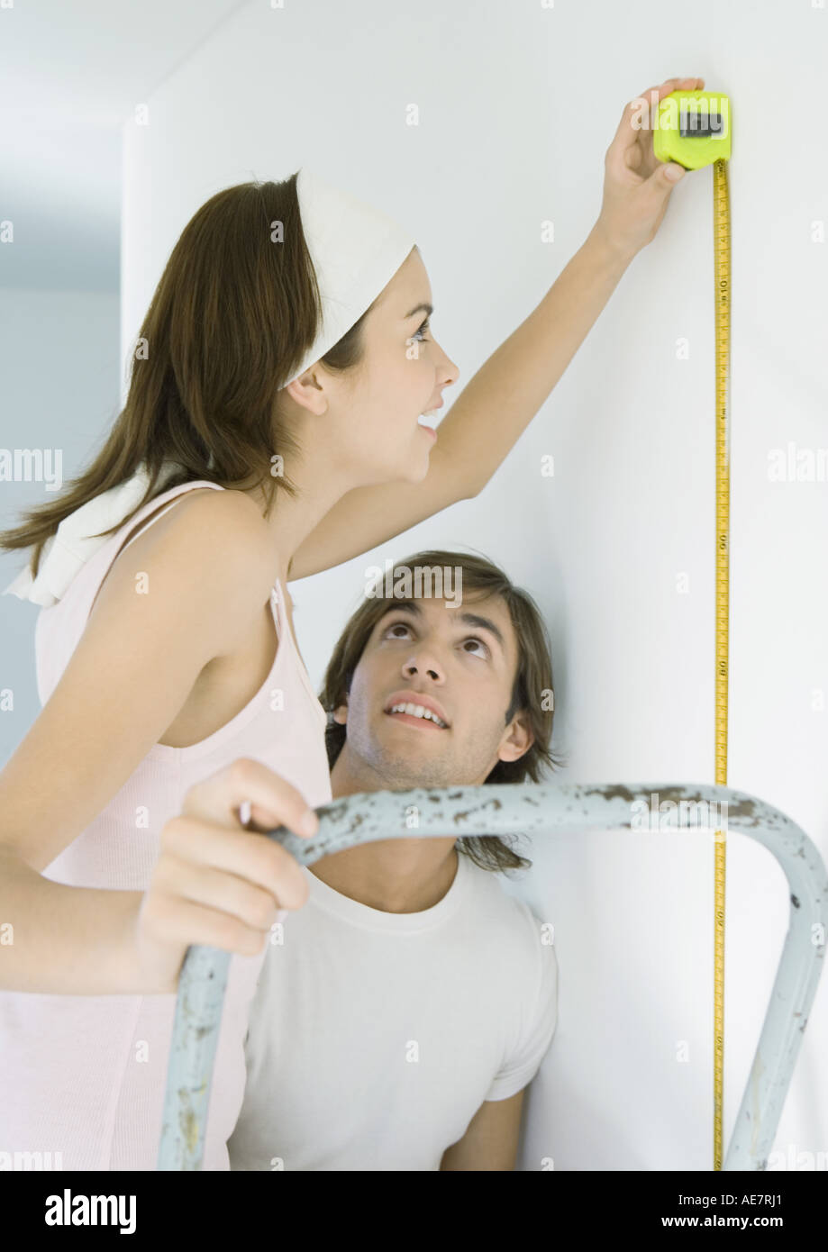 Woman on ladder using measuring tape as man watches Stock Photo