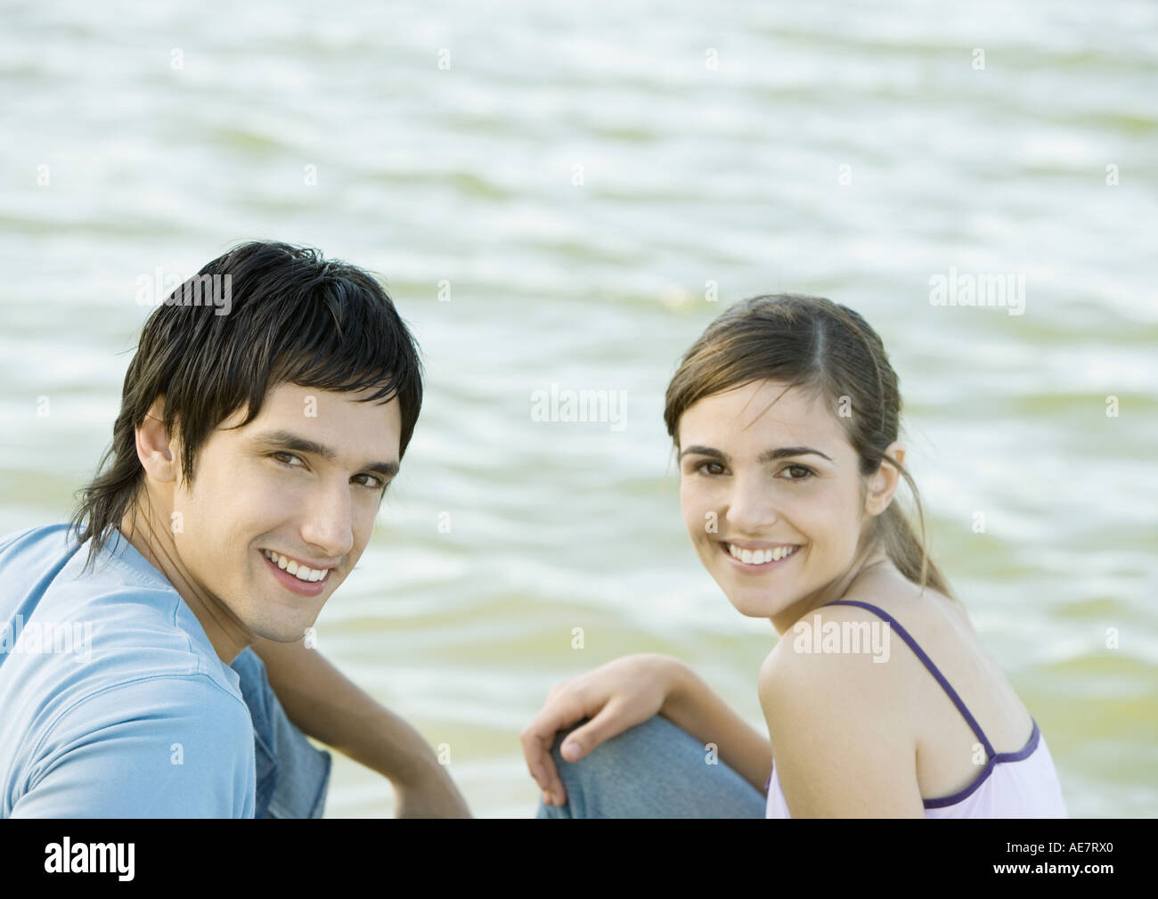 Young couple sitting side by body of water, smiling and looking at camera Stock Photo