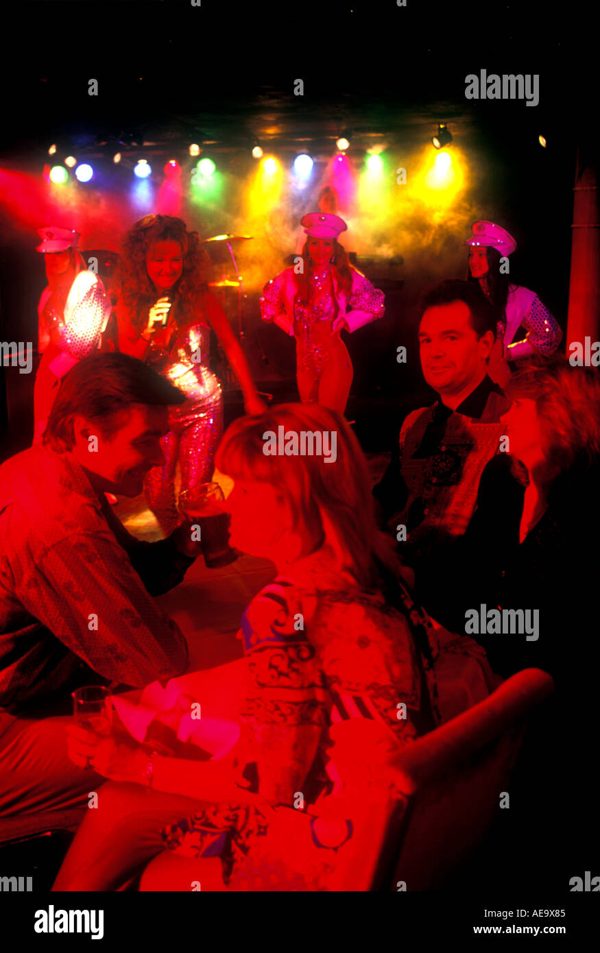 two 2 couples sitting down in disco night club with very red lighting on faces enjoying dancers dancing Stock Photo