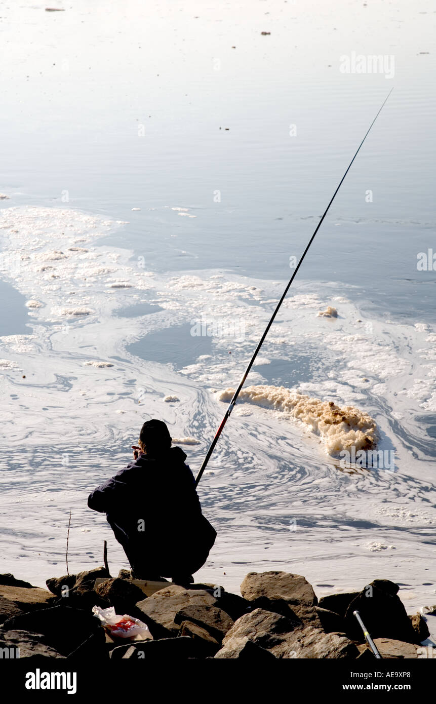 A man fishing in a dirty river water filled with foam and peat residue from a power plant , Finland Stock Photo
