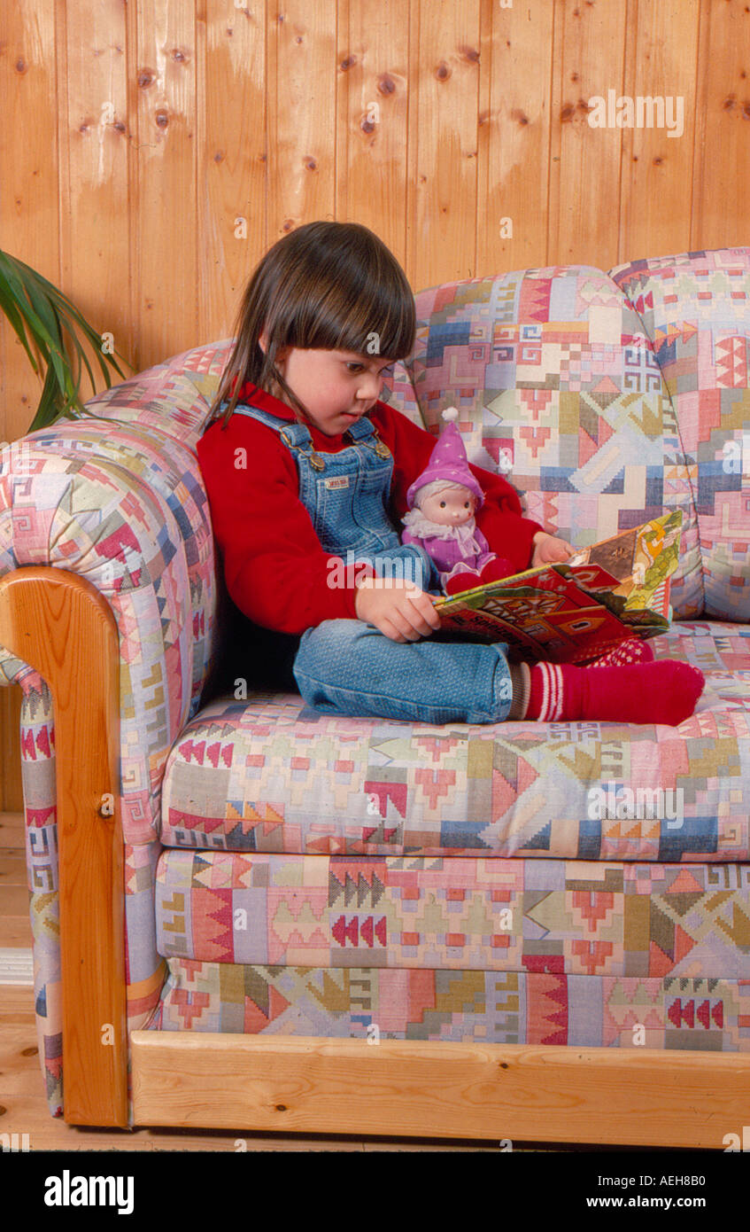 girl sitting with reading book and doll on sofa. Photo by Willy Matheisl Stock Photo