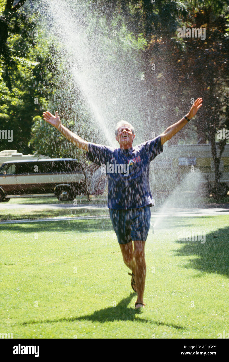 Mature man running through sprinklers water playing arms stretched out loves life freedom abandonment careless worry free childlike playful Myrleen Pearson Stock Photo