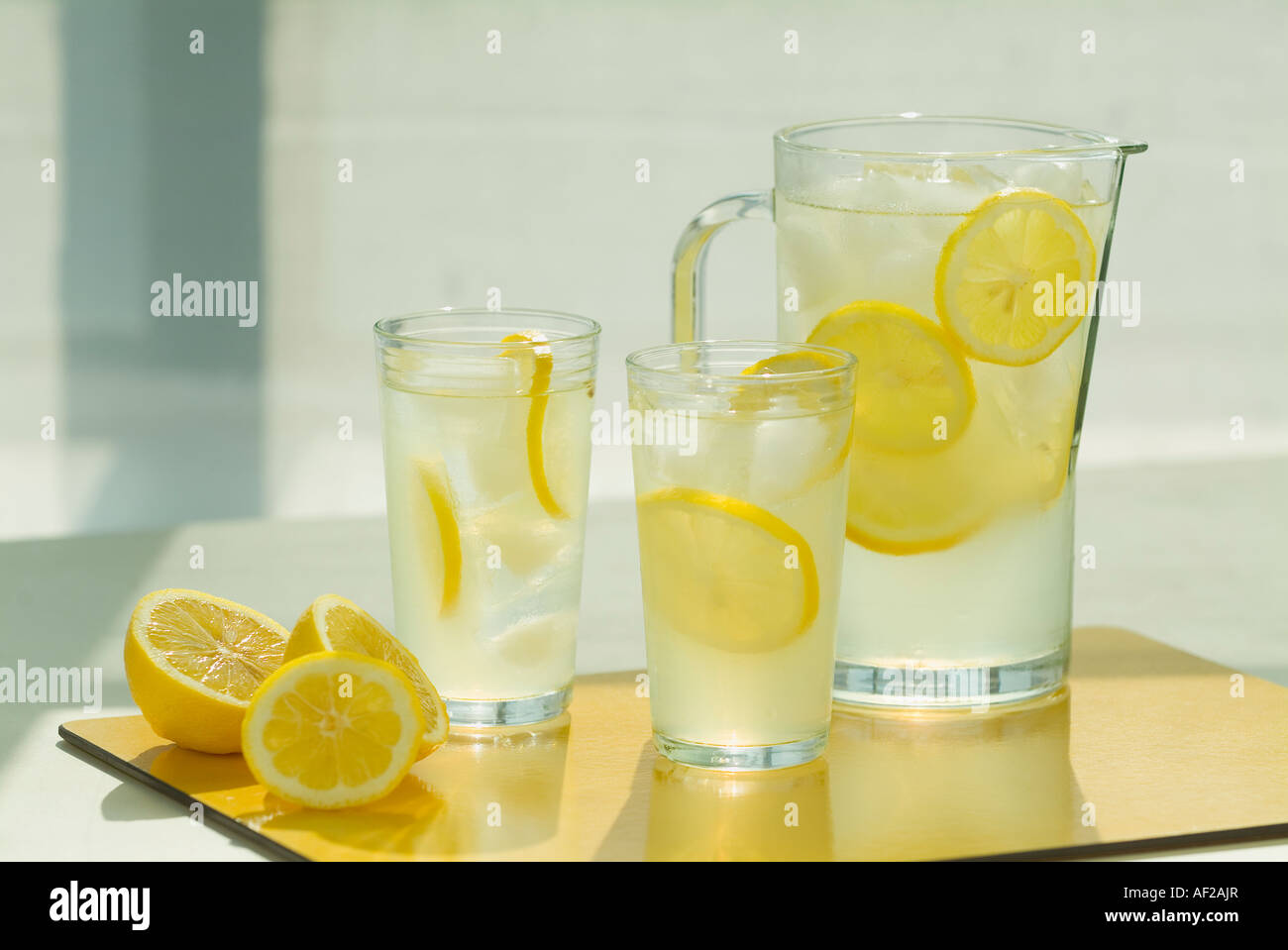 A sunlit pitcher and glasses full of fresh lemonade, bathed in sunlight and  surrounded by cut lemon slices. Stock Photo