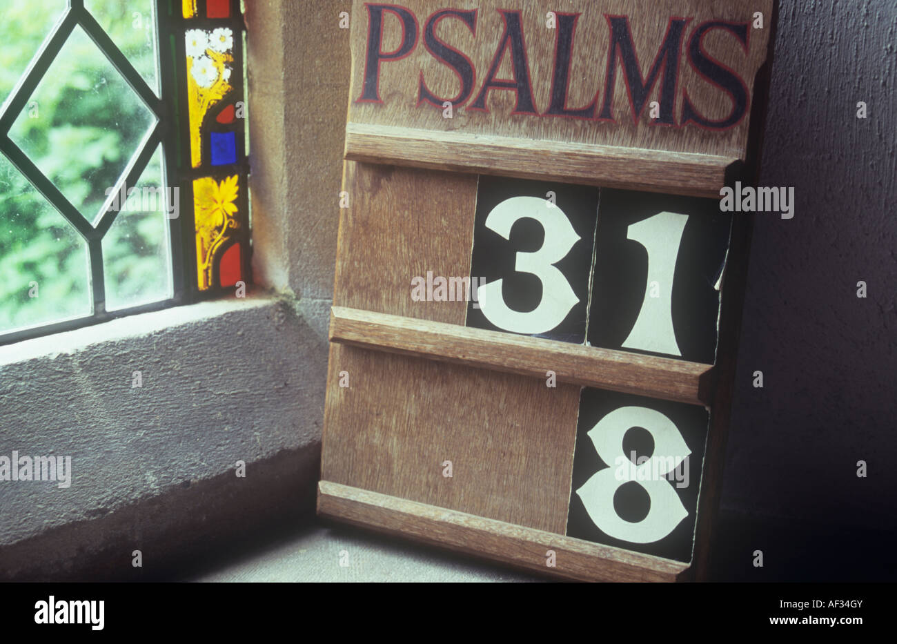 Board indicating selected Psalm numbers on sill next to diamond leaded window with stained glass border and greenery outside Stock Photo
