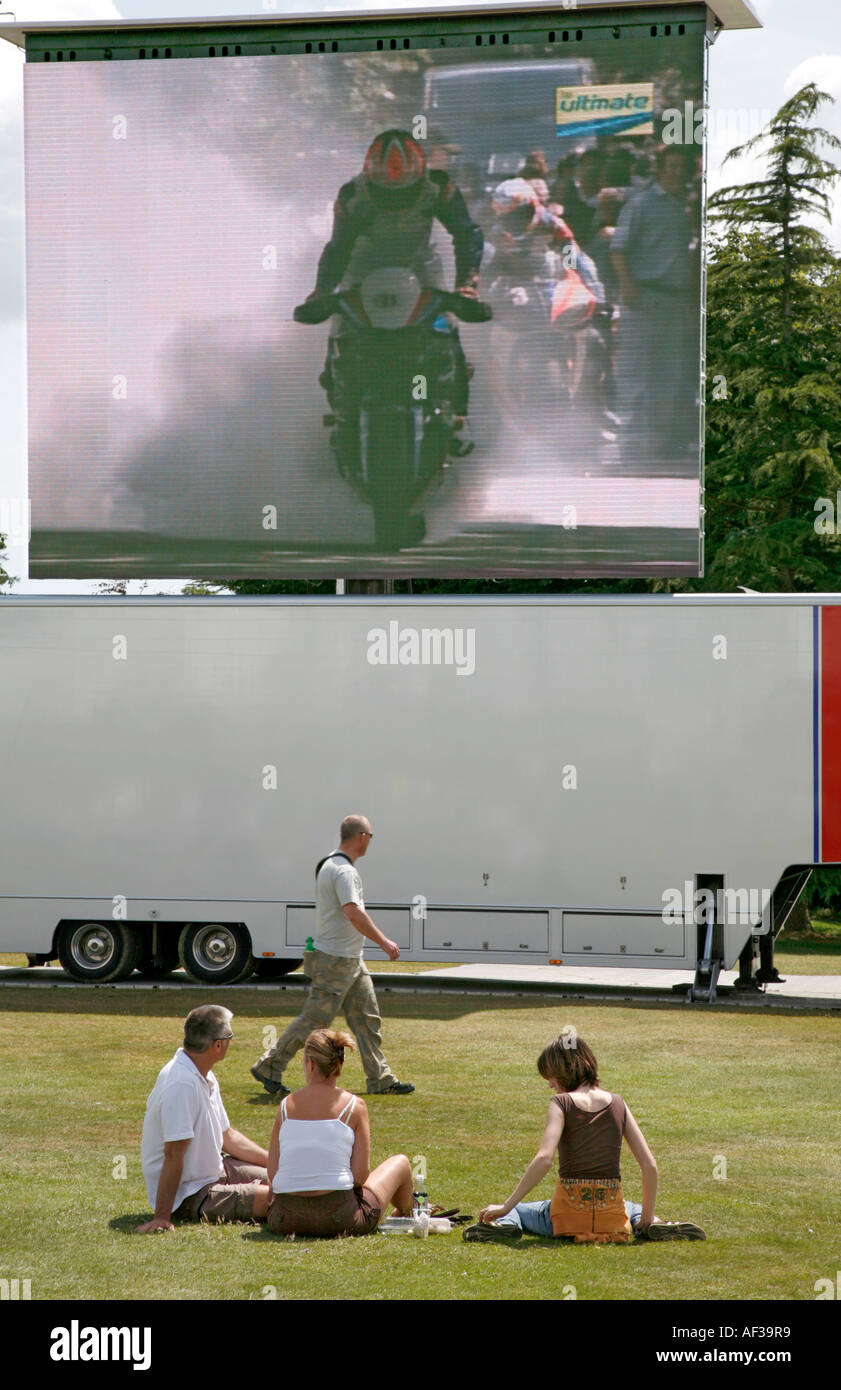 Spectators sitting on the grassed area in front of giant TV at the Goodwood Festival of Speed. Stock Photo