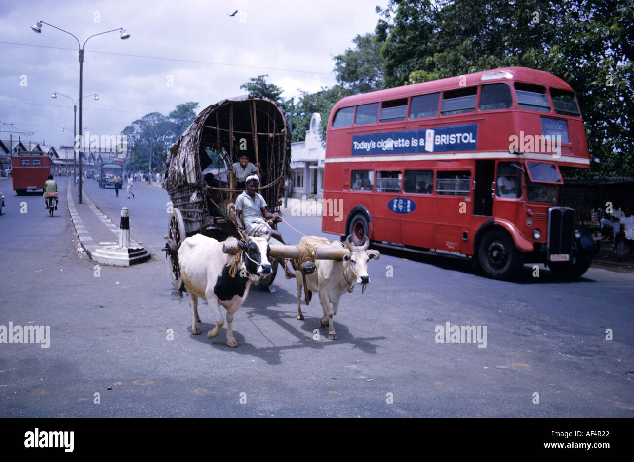 Old ox drawn cart and bright red ex London bus with Bristol cigarette advert on the side in mid 1960s in Colombo Sri Lanka Stock Photo