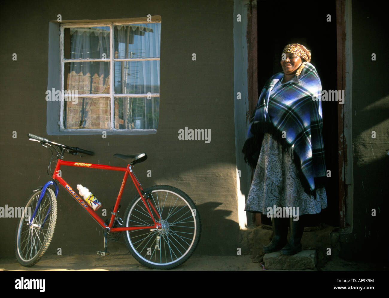 Black woman standing in the doorway of her homestead, next to a bright red mountain bike. Eastern Free State; South Africa Stock Photo