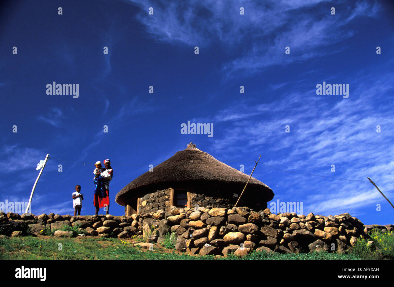 A woman standing outside her neat stone and thatch homestead with her children. Lesotho Stock Photo