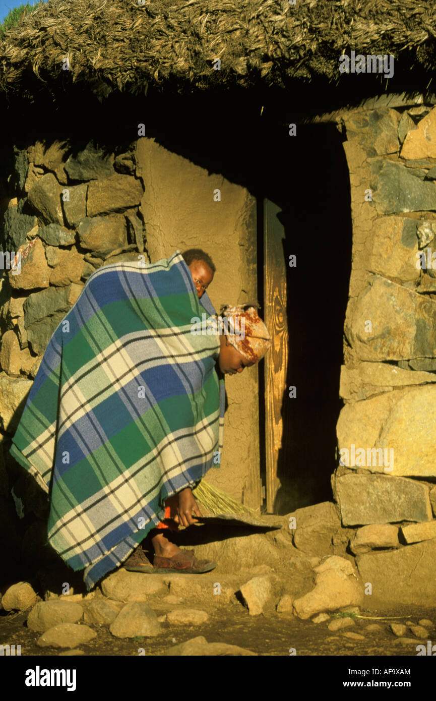 Woman with a baby tied onto her back with a blanket, sweeping the steps of her stone and thatch hut. Stock Photo
