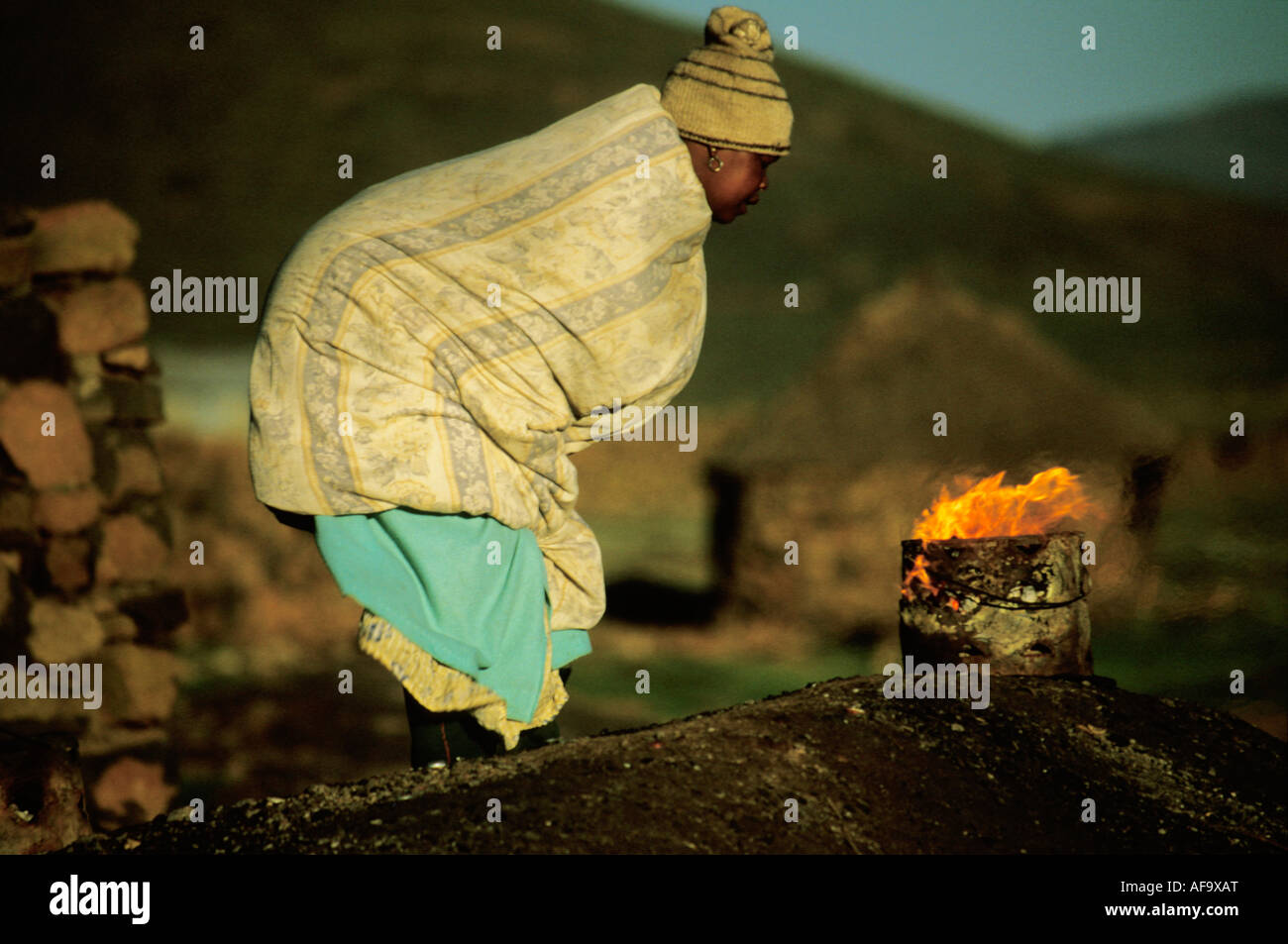 Woman with blanket wrapped around herself leaning over a small fire