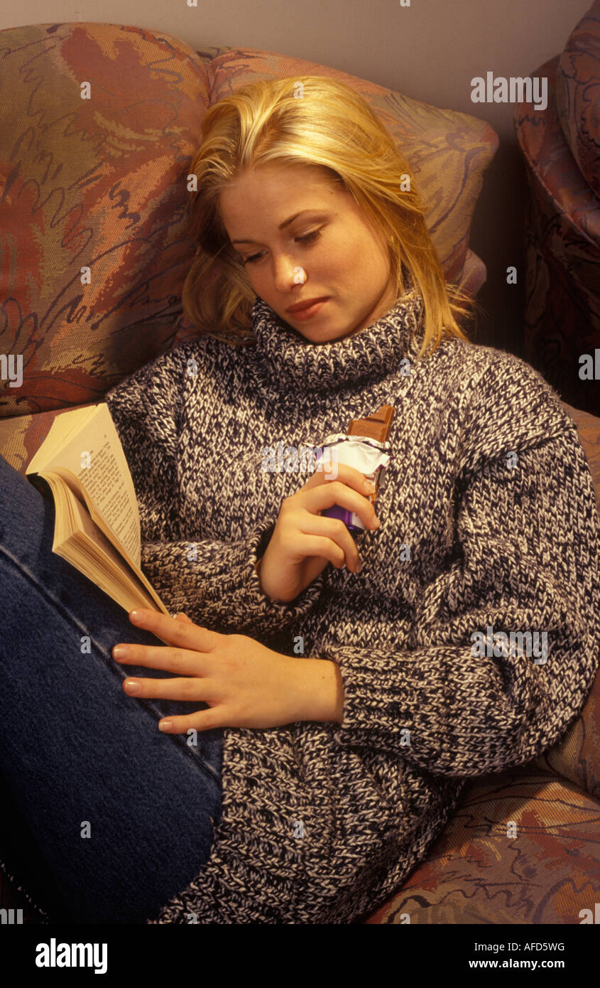 woman curled up with a book eating a bar of chocolate Stock Photo