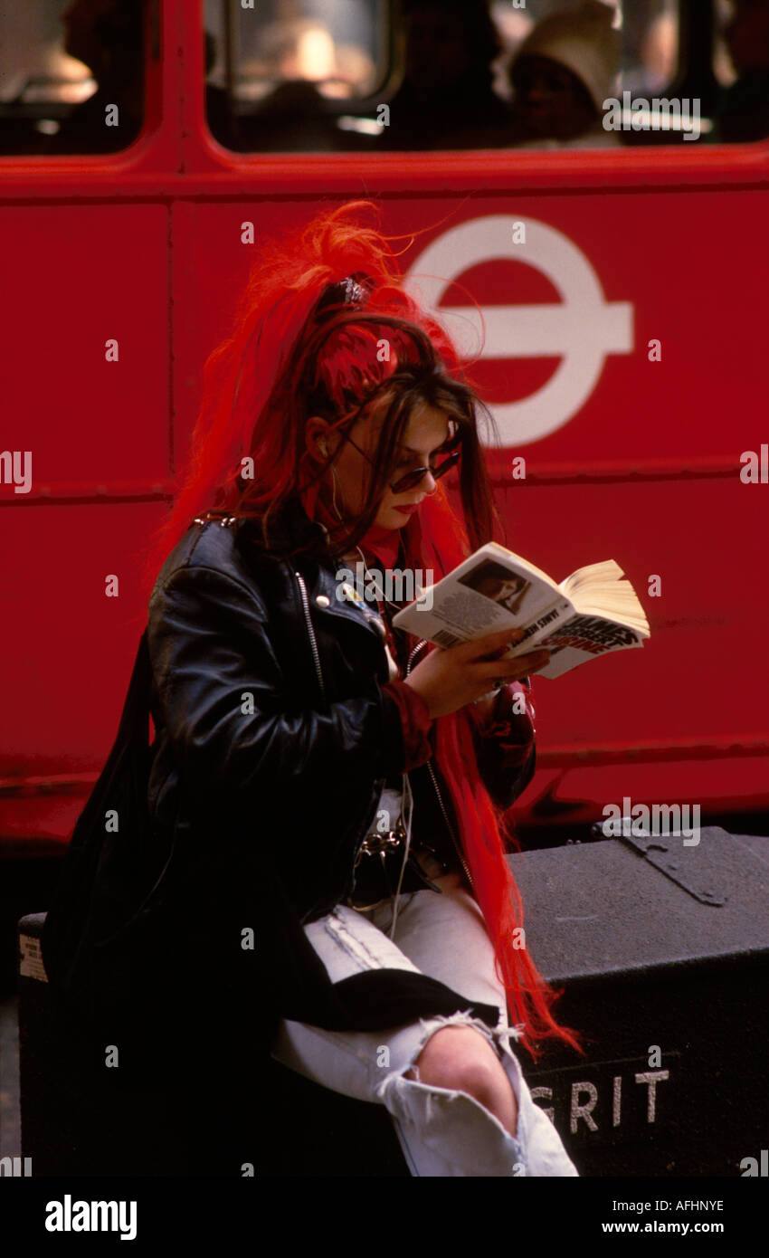 A young punk girl with bright red hair extensions sits on a road gritting box on a London street reading as a bus drives past. Stock Photo