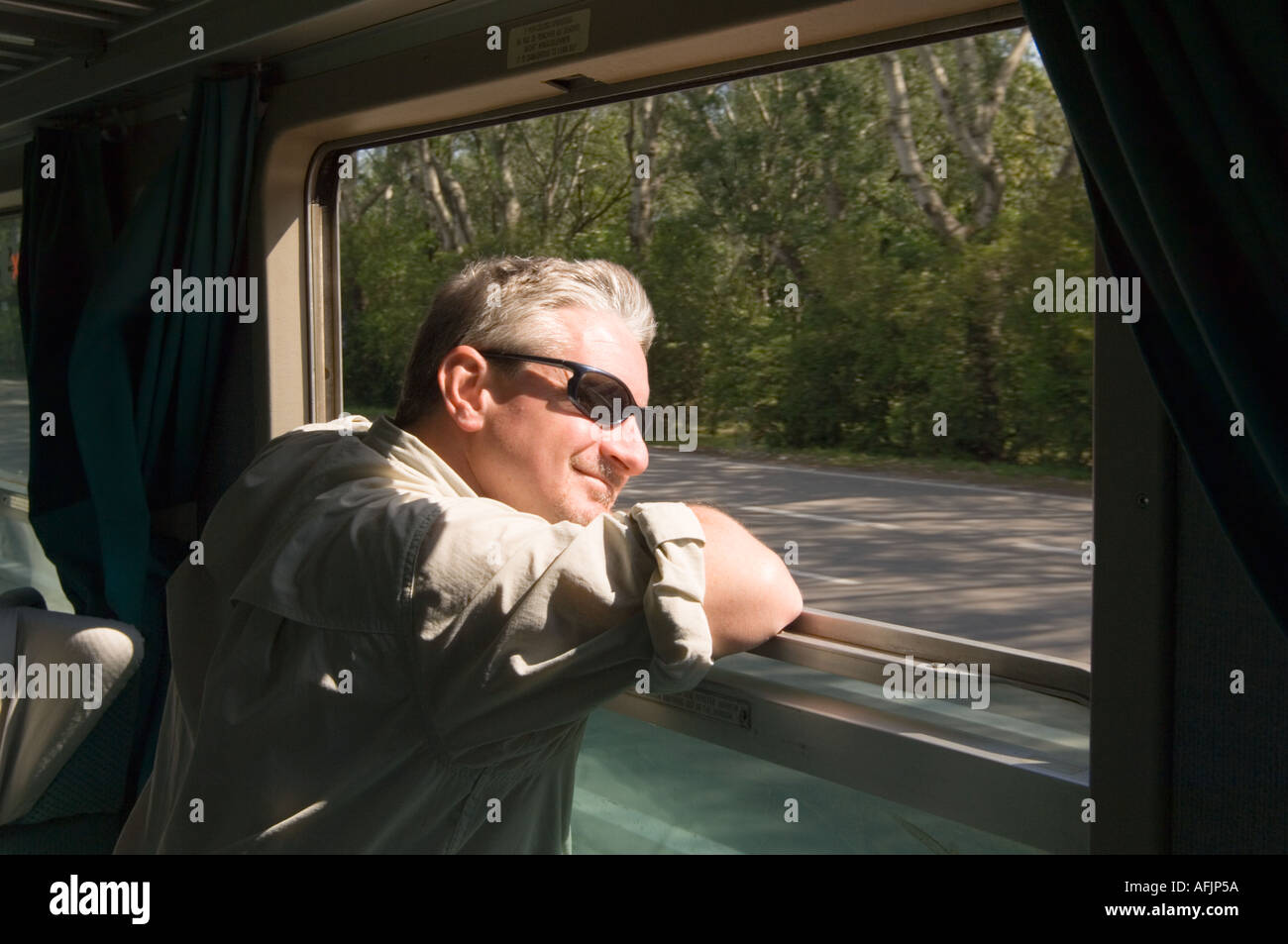 middle aged man in a travelling shirt and sunglasses leaning on his elbows out the window of Italian train car with sun on face Stock Photo