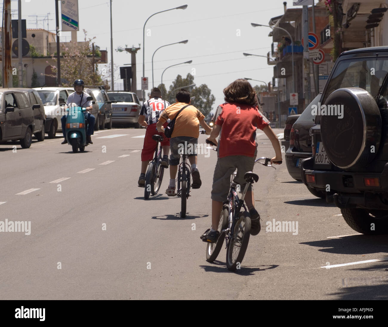 three kids on bikes riding away from viewer and one man on a scooter riding toward viewer on a street on Sicily in Italy Stock Photo