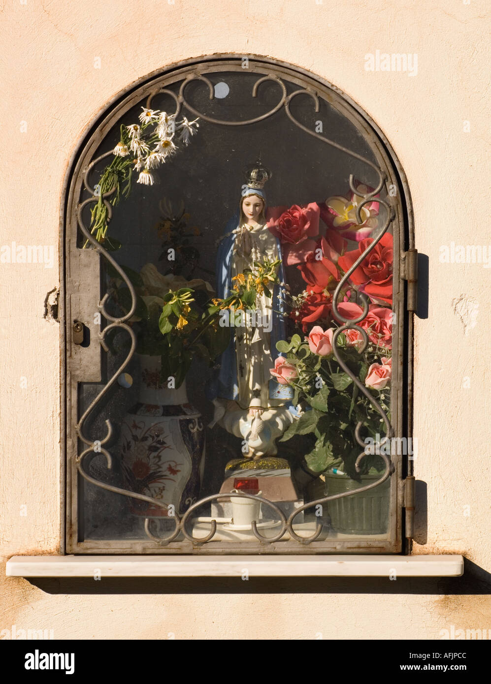 sunlit shrine to the virgin Mary set in an alcove in a stucco wall with flower offerings and a glass door with wrought iron Stock Photo