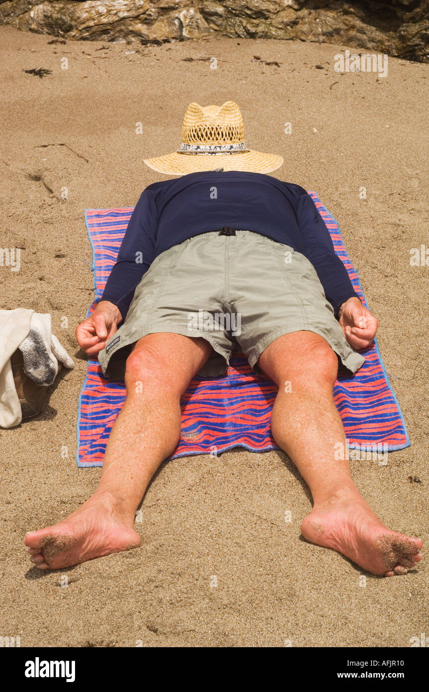 man lying on a towel on a beach with a straw hat covering his face barefoot in shorts and a long sleeved shirt Stock Photo