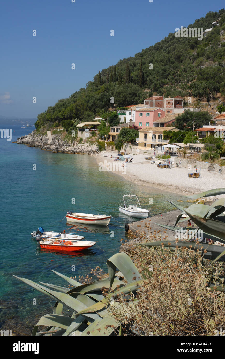 Small beach in Nissaki bay, Corfu, Greece with four hire boats and cacti in foreground, buildings and tavernas beyond Stock Photo
